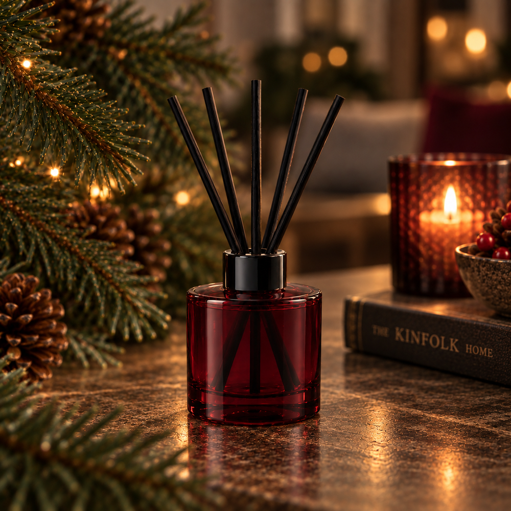 A red glass reed diffuser bottle with black reeds stands on a tabletop in a warm holiday setting, surrounded by pine branches, a lit candle, and a book.