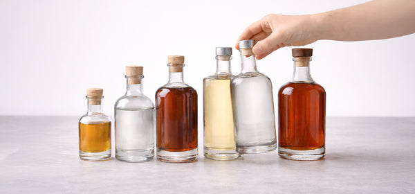 A row of clear glass spirit bottles in different sizes with cork stoppers, filled with amber and clear spirits