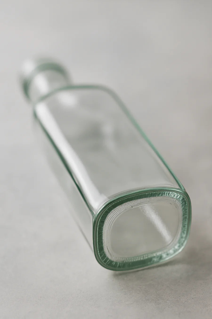 An empty, clear glass bottle with a stopper, lying on its side on a light gray textured surface.