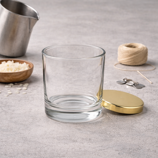 Clear straight-sided candle glass with a gold lid, beside soy wax flakes, cotton wick and wick sustainers
