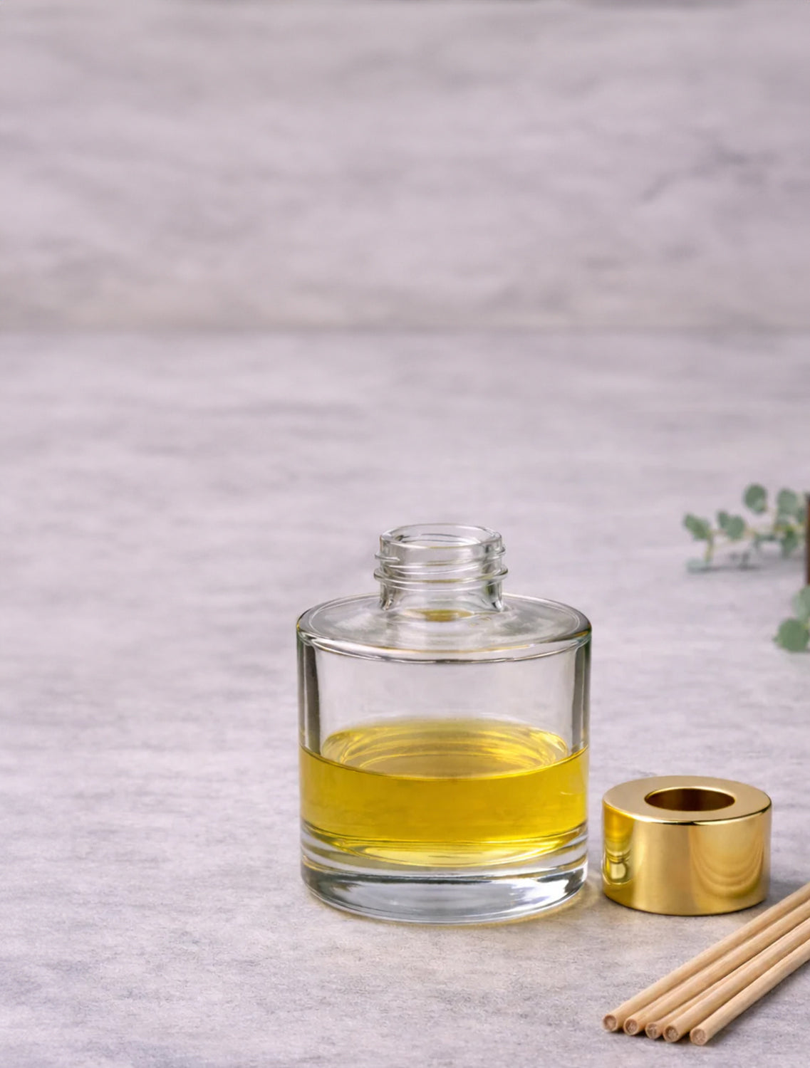 Clear glass bottle with yellow liquid, gold candle holder, and wooden sticks on a light gray background