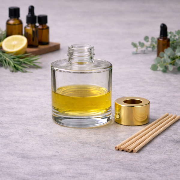 Clear round glass diffuser bottle filled with fragrance oil, with a gold diffuser cap and natural reeds beside it