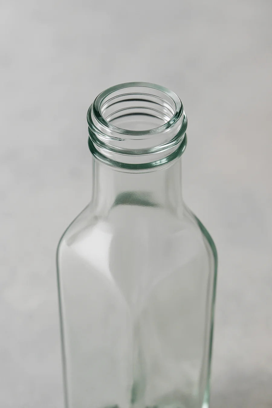 An empty clear glass bottle with a threaded neck, shown from a high-angle perspective against a neutral light gray background.