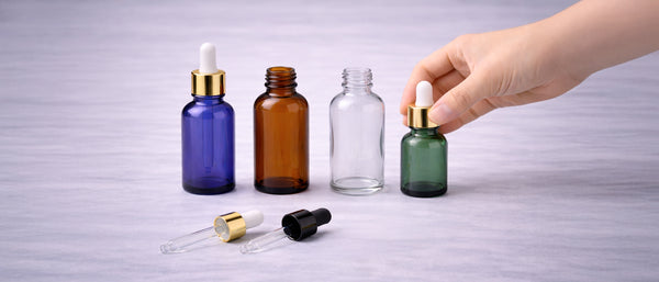Blue, amber, clear and green glass dropper bottles in a row, with a hand placing a gold pipette cap on the green bottle and two loose pipettes in front